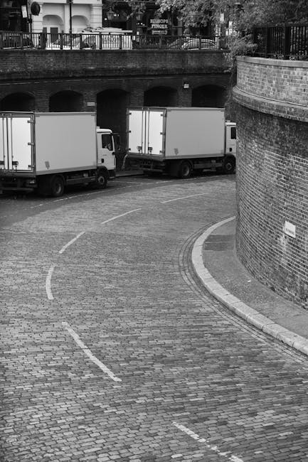 A black-and-white photograph showing a curved cobbled driveway leading into an underground garage or loading area underneath a residential building. Two white standard box trucks are parked, with thei