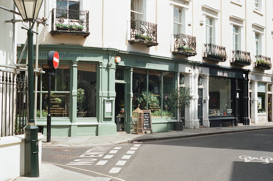 Photograph of a street view in Kensington showing a light green shopfront with large glass windows and a sign that reads 'Kensington Movers' indicating a moving company. The shop is situated inside a white, multi-story building with five small balconies with black wrought iron railings and potted plants. A black chalkboard sign is placed outside, and potted plants and greenery are visible through the windows. To the left, a metal gate and a no-entry traffic sign are present, along with a small bollard and a bike lane marking on the asphalt. The scene captures elements of home relocation logistics, including the storefront potentially used for packing and moving services, with lighting suggesting daytime conditions.