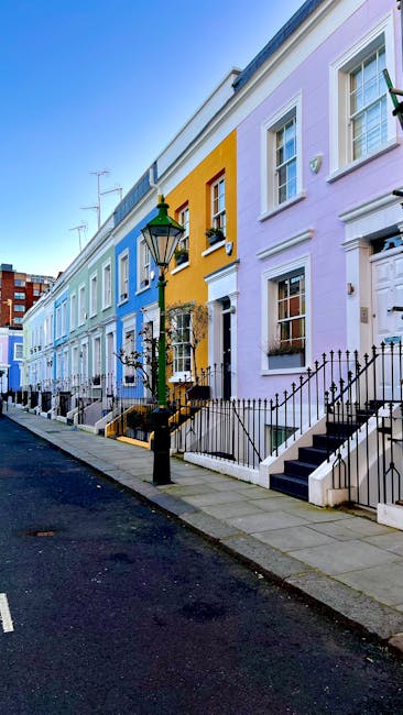 A row of colorful terraced houses in Kensington, featuring facades painted in pastel shades of blue, yellow, and purple, with white-framed windows and black wrought-iron railings along the front steps. The pavement is clean and lined with a black lamppost and small decorative trees in planters, set against a bright, clear sky. The scene depicts a typical urban residential street in Kensington, demonstrating the area's conservation character. The image is part of a visual illustration related to house removals and moving logistics, highlighting the exterior environment where furniture transport and home relocation processes take place, with visible stairs leading to front doors and a smooth sidewalk surface often encountered during packing and loading activities by professional movers such as Kensington Movers.