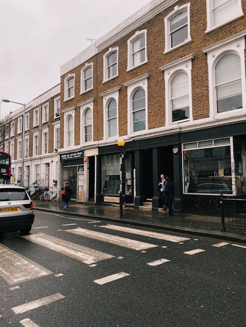 The image depicts a street scene outside a multi-story brick building in Kensington on a rainy day, with a black shopfront on the ground floor and several large windows on the upper floors. A black van is parked on the wet road, partially visible on the left side, with a white car behind it. Two pedestrians, one wearing a dark coat and the other a red jacket, are walking on the sidewalk near the shopfront. A person dressed in dark clothing and a face mask is standing near the entrance of the building, possibly involved in the home relocation process. The scene includes a yellow streetlamp and a bicycle parked against the building's wall. The weather appears overcast, with reflections on the wet pavement, indicative of recent or ongoing rain. This setting suggests a typical urban environment suitable for household removals, with potential access issues for large furniture or boxes during the loading process that Kensington Movers might manage as part of their removals services in the area.