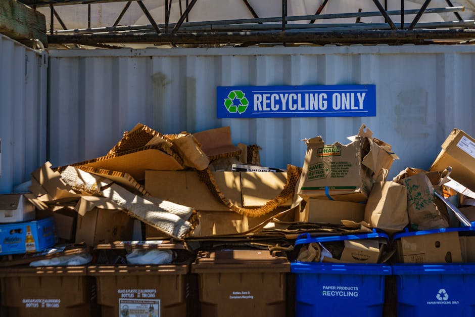 Close-up image of a large pile of crushed and crumpled aluminium cans and plastic bottles, primarily recyclables such as soda cans and beverage containers, scattered on the ground. The cans display va