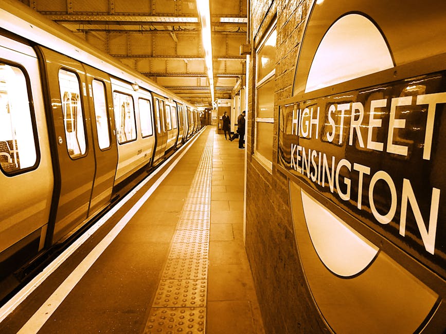 Inside the underground platform at High Street Kensington station, the image shows a yellow and white London Underground train parked along the platform with its doors closed. The platform features a tactile paving strip for visually impaired travelers running parallel to the train, and a brick wall with a large, illuminated roundel sign displaying the station name, 'High Street Kensington,' mounted on the wall on the right. Several passengers are visible standing on the platform, some closer to the train and others near the sign, waiting to board or disembark. The station's ceiling is lined with lighting strips that illuminate the area evenly, and the overall scene depicts a typical subway station environment suitable for house removals or relocation services, such as those provided by Kensington Movers, where careful planning and transportation logistics are essential for handling furniture and belongings in Victorian properties or similar residential settings.