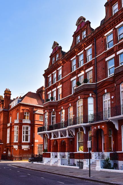 Photograph depicting the exterior of a red brick Victorian residential building on Holland Park Road in Kensington. The building features multiple large sash windows with white frames, decorative iron balconies on the first-floor windows, and a white wooden canopy supported by brackets over the entrance steps. The pavement in front of the property shows a stack of packed cardboard moving boxes, some wrapped in plastic or fabric protective covers, and a hand truck or trolley is being used to transport these boxes. Nearby, additional cardboard cartons and wrapping materials are arranged on the ground as part of a household packing process. Visible in the background are other similar Victorian buildings with ornate architectural details, under a clear blue sky. This scene illustrates the initial stages of a home relocation involving furniture transport and packing preparatory to moving day, as managed by Kensington Movers, a professional removals company specializing in house moves within historic Victorian properties.