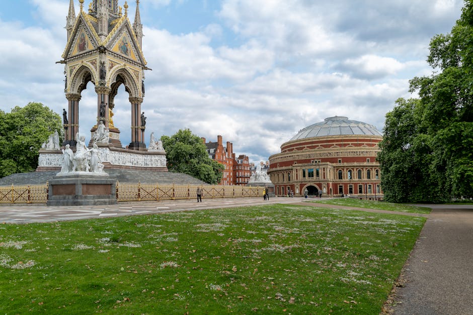 A city park scene featuring a large, ornate monument with multiple statues and decorative elements made of stone and metal, situated on a paved area with a low fence around it. To the right, there is a historic round brick building with a domed roof, surrounded by green trees and grass, with a pathway leading past the building. The sky above is partly cloudy with patches of blue visible. The area appears calm with a few pedestrians in the distance, suggesting a typical urban park environment. This setting illustrates a location suitable for exterior house removals or furniture transport as part of a professional home relocation or moving service by Kensington Movers.