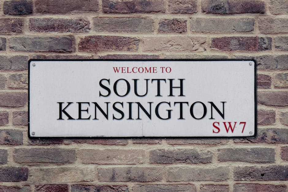 A rectangular metal street sign mounted on a brick wall displaying the message 'Welcome to South Kensington SW7'. The sign has a white background with black and red text, with 'South Kensington' prominently centered in large black letters, and smaller red and black text indicating the area's postal code. The brick wall behind the sign is composed of reddish-brown bricks arranged in a horizontal pattern, with some bricks showing variations in texture and color. The sign appears to be part of a home relocation or moving process, as it is situated outside a building in the South Kensington area, potentially indicating the start or destination point for furniture transport or packing and moving services provided by Kensington Movers.