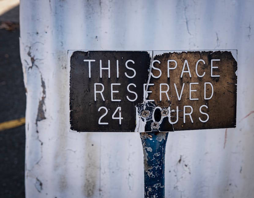 A close-up image of a weathered metal parking sign mounted on a blue-painted pole, with a black and white background wall behind it. The sign reads 'THIS SPACE RESERVED 24 HOURS' in white capital letters, with visible rust, peeling paint, and scratches on its surface. The sign appears to be attached by screws and has some damage, including a missing section and torn areas near the middle. The surrounding environment suggests an urban setting, possibly near a residential building or a loading zone, reflecting typical parking restrictions in Kensington. This image relates to parking regulations relevant to house removals and loading procedures, highlighting the need for proper permits during home relocation and furniture transport, as managed by Kensington Movers.