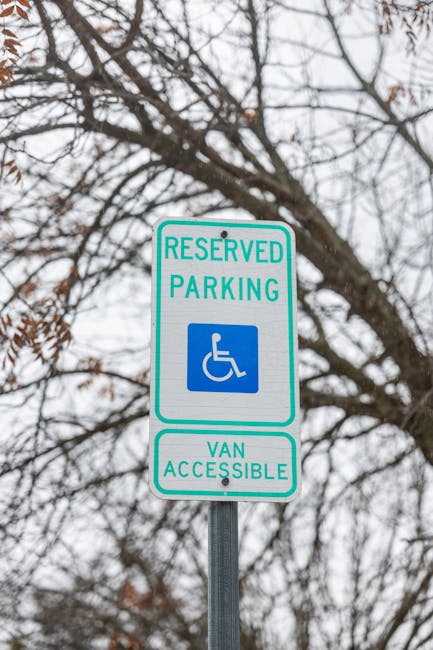 A close-up view of a white reserved parking sign with green borders, mounted on a metal pole beneath a tree with bare branches. The sign features a blue wheelchair symbol indicating accessibility for disabled parking, with the text 'RESERVED PARKING' above and 'VAN ACCESSIBLE' below. The background shows an overcast sky and tree branches without leaves, suggesting a winter or late autumn setting. In the context of house removals and moving logistics, such parking restrictions are relevant for planning loading and unloading zones, which companies like Kensington Movers must consider during home relocation processes in areas like Kensington.