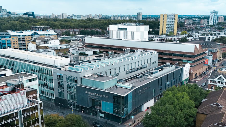 An aerial view of the Royal College of Art building, a modern structure with a combination of glass and metal panels, situated among other commercial and residential buildings in the surrounding urban area. The rooftop features skylights and ventilation units, with some construction or maintenance equipment visible. Adjacent streets include pavement and green trees, with a few parked vehicles along the roadside. The environment appears well-lit with natural daylight, highlighting the nearby cityscape consisting of high-rise apartment blocks and office buildings in the distance. This setting reflects an urban context suitable for house removals and furniture transport services provided by Kensington Movers, who assist with packing, loading, and home relocation logistics within the borough of Kensington and Chelsea.