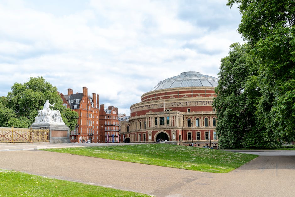 A wide view of the Royal Albert Hall, a large circular building with a domed roof made of grey metal, situated in a park area with green grass and pathways. To the left of the hall, there is a white marble statue featuring multiple figures, placed on a raised pedestal. Surrounding the hall and statue are trees with lush green foliage, and in the background, there are red-brick residential buildings with multiple chimneys. The scene is captured during daylight hours with partly cloudy skies overhead. No furniture or packaging materials are visible in this image. The setting appears to be tranquil, suitable for house removals or furniture transport preparation, with the open space and accessible pathways evident. Kensington Movers could potentially utilize this space for loading or unloading vehicles during home relocation or packing and moving services.