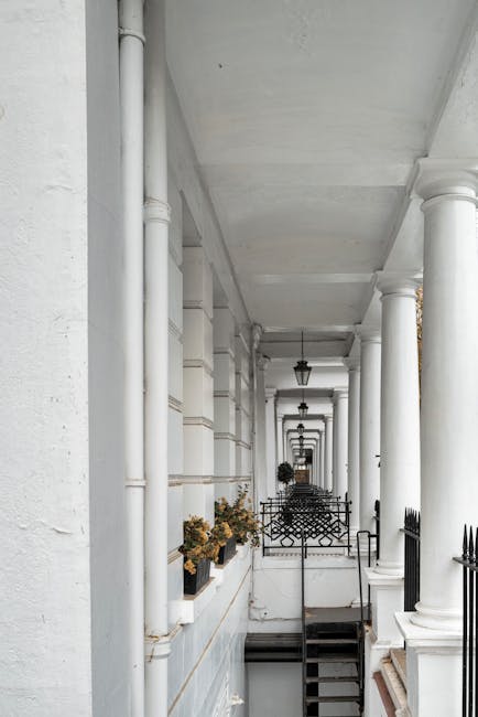 A long, narrow outdoor corridor showcasing a row of white columns supporting a ceiling with white paint and decorative detailing. The corridor's left side features a white wall with built-in window boxes filled with dried plants, and black iron railings are visible along the edge. Several black vintage-style lanterns hang overhead, evenly spaced along the corridor. The floor appears to be made of dark stone or concrete, and a set of small steps is visible toward the far end. There are no visible furniture or objects related to removals or moving activities, suggesting the area is part of a residential building's exterior walkway arranged with classic design elements, suitable for house removals or relocation services near South Kensington.