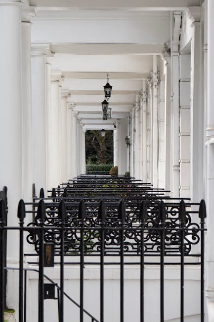 A covered porch area of a residential property featuring white-painted wooden columns and a ceiling, with ornate black wrought iron railings and gates at the front. Hanging lantern-style light fixtures are suspended from the ceiling. Beyond the porch, there is a garden with a large tree and greenery visible through the open space. The environment is well-lit with natural daylight, and the setting appears to be prepared for house removal or furniture transport, with the porch acting as a staging area for loading or moving process. Kensington Movers' team could utilize this space for packing, moving furniture, or loading items into vans, relating to house removals and relocation services near South Kensington station.